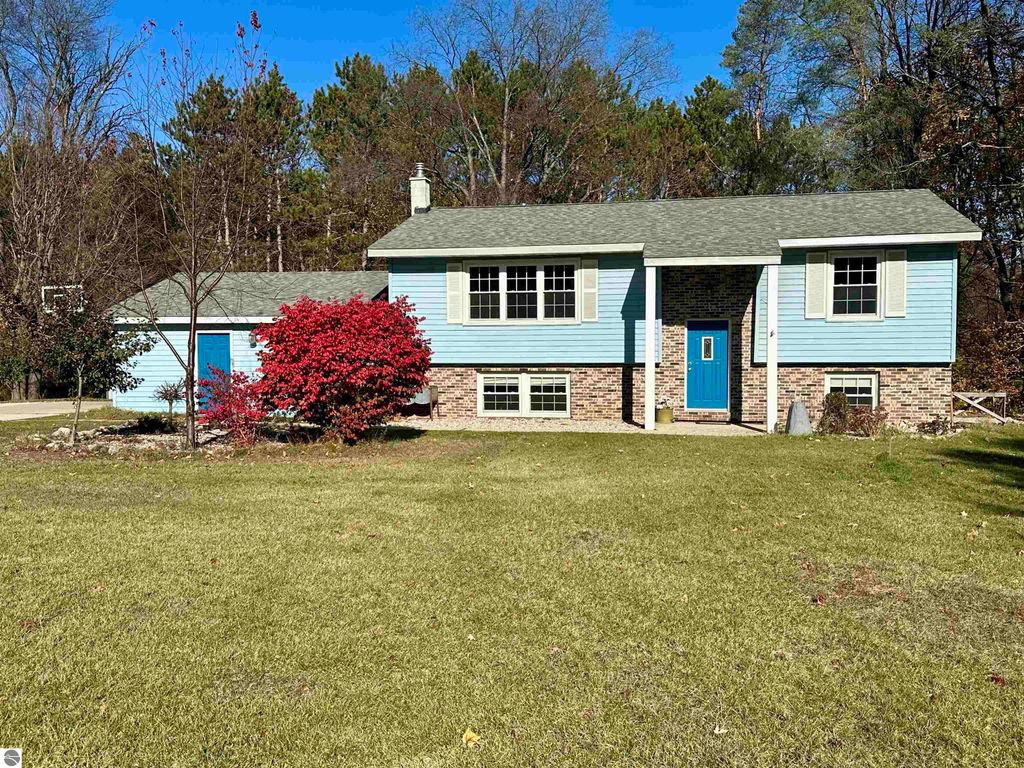 Bi-level home at 8398 W Whispering Pines, Lake City, MI, featuring blue siding, brick accents, and a vibrant red bush in the front yard, set in a peaceful wooded area.