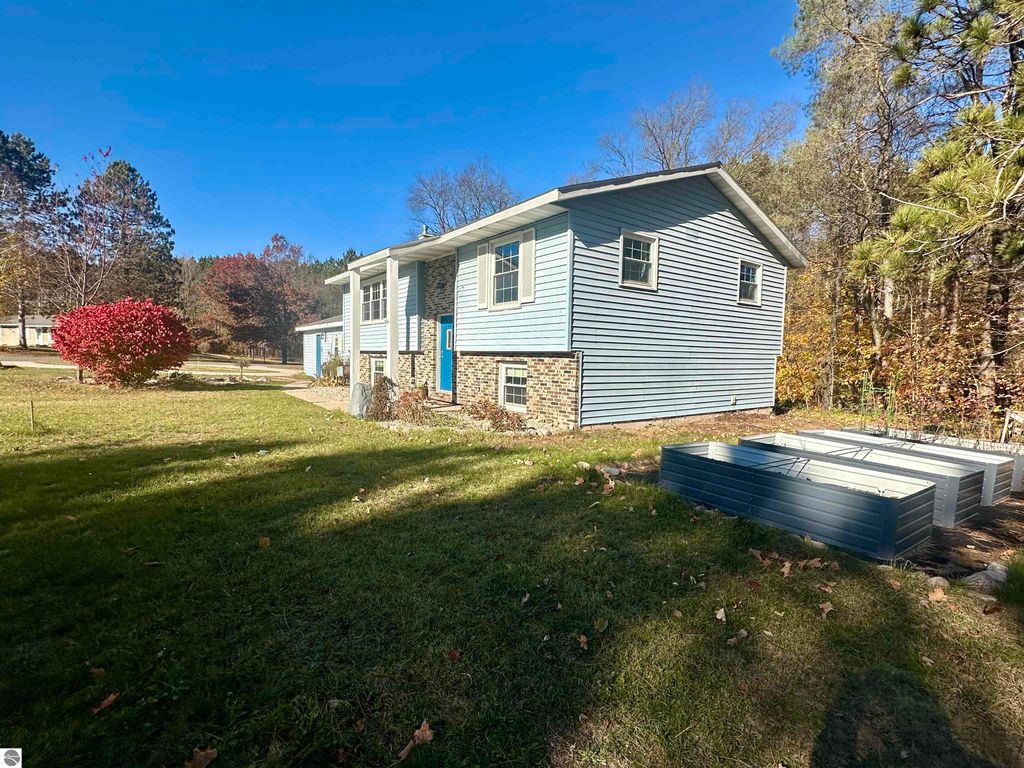 Bi-level home at 8398 W Whispering Pines, Lake City, MI, featuring a blue exterior, landscaped yard, and nearby garden beds, set in a quiet subdivision surrounded by trees.