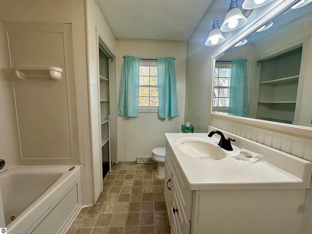 Bathroom interior featuring a white vanity with sink, modern faucet, and mirror, light fixtures above, bathtub on the left, window with light blue curtains, and beige tiled floor, showcasing the home's inviting design at 8398 W Whispering Pines, Lake City, MI.