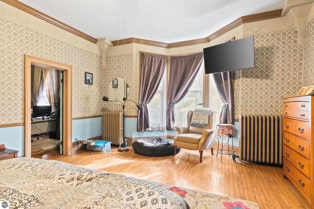 Victorian bedroom with original wood trim, high ceilings, and modern amenities, featuring a cozy reading nook, decorative curtains, and a flat-screen TV, reflecting the charm of the historic home at 8195 Fifth Street, Onekama, MI.