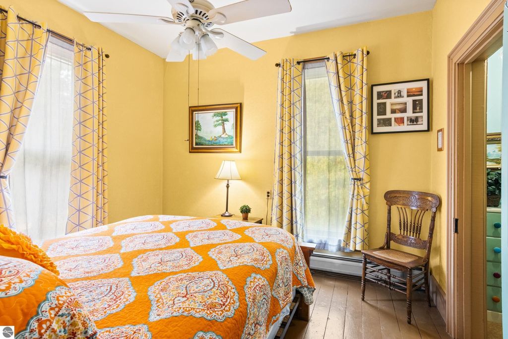 Brightly colored bedroom featuring a yellow wall, patterned orange quilt, large windows with decorative curtains, a bedside lamp, and a vintage wooden chair, showcasing the charming interior of the Victorian home listed at 8195 Fifth Street, Onekama, MI.