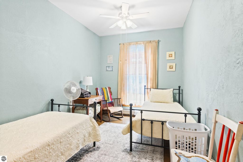 Victorian-style bedroom with two metal beds, light blue walls, a ceiling fan, and natural light from a large window, featuring a cozy chair and decorative elements, ideal for a bed and breakfast setting.
