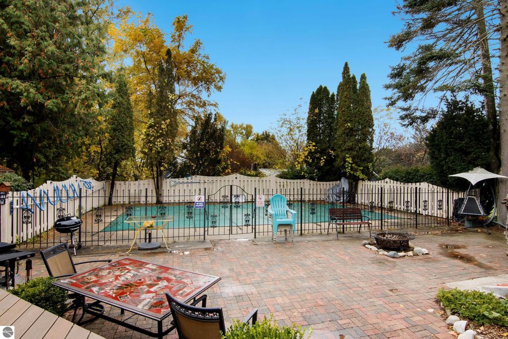 Backyard view of a Victorian home in Onekama, featuring a fenced in-ground pool, patio area with a decorative table, and lush greenery, highlighting the property's charm and outdoor amenities.