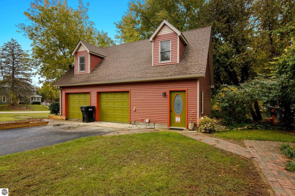 Victorian-style home exterior at 8195 Fifth Street, Onekama, MI, featuring a pink facade, green garage doors, and a decorative front door, surrounded by lush greenery and a paved walkway.