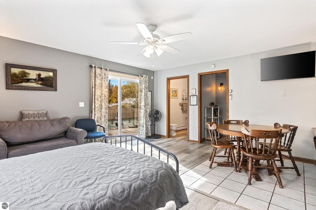 Cozy interior of a Victorian-style bedroom with a queen bed, seating area, and dining table, featuring a ceiling fan, wall-mounted TV, and large windows overlooking the backyard pool area.