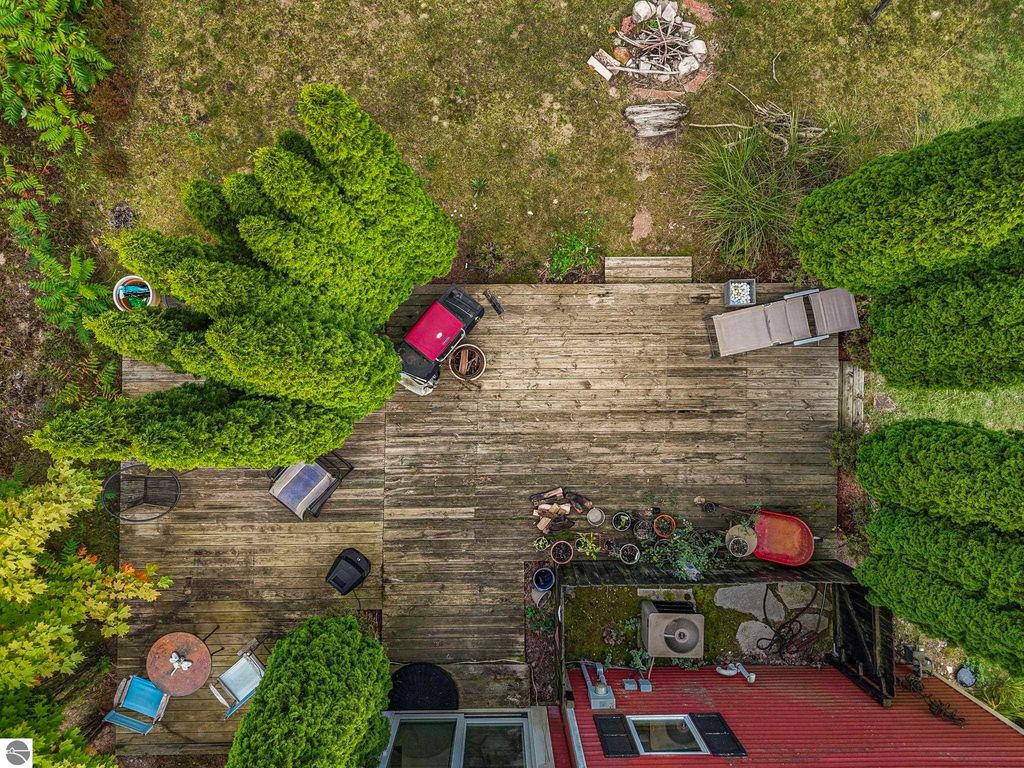 Aerial view of a wooden deck surrounded by lush greenery, featuring outdoor furniture, a fire pit, and potted plants, near a farmhouse on 3 acres in Leland, MI.