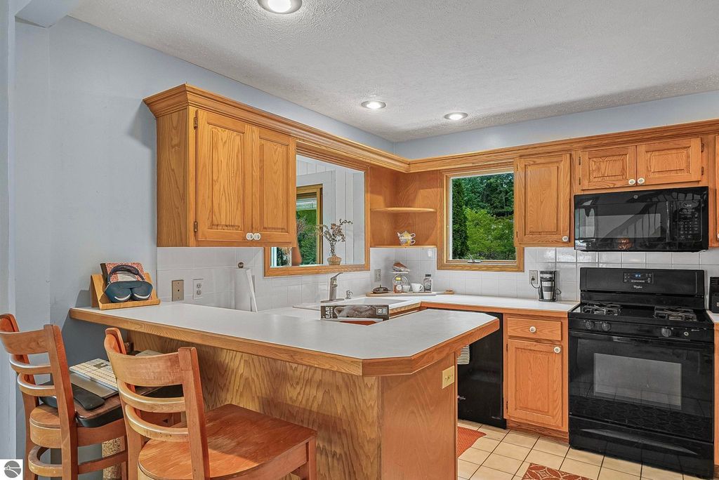 Kitchen interior of elevated farmhouse at 5361 N Manitou Trail, featuring wooden cabinets, black appliances, and a breakfast bar, with a window showing greenery outside.