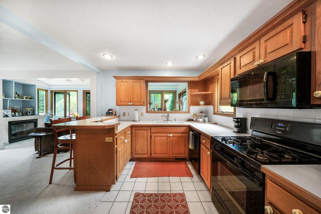 Kitchen interior of a farmhouse at 5361 N Manitou Trail, featuring wooden cabinets, gas stove, and breakfast bar, with views of surrounding greenery through large windows.
