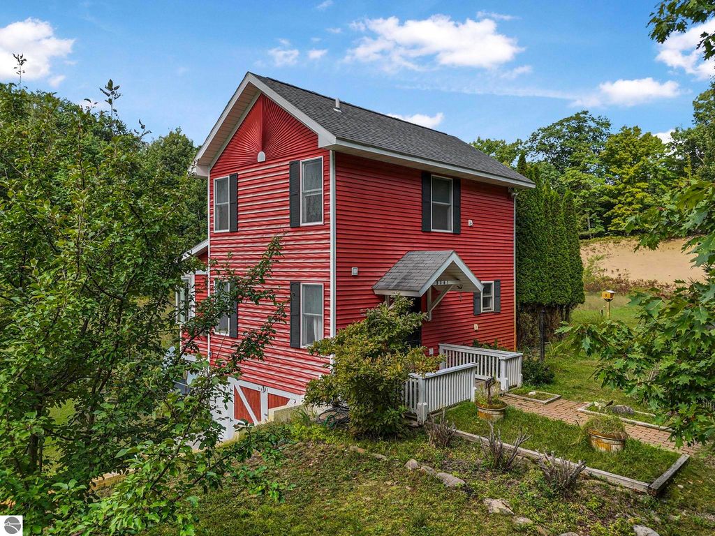 Elevated red farmhouse surrounded by greenery, featuring a front porch and stone pathway, located on 3 acres near Leland, MI.