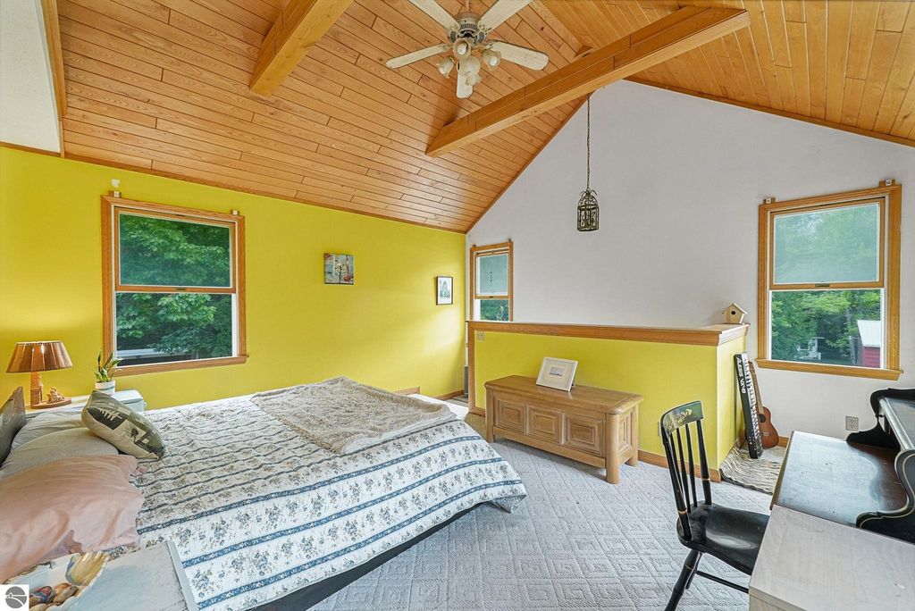 Bright and airy primary bedroom with vaulted wood ceiling, yellow walls, large windows, and cozy furnishings, showcasing a comfortable bed and a workspace, located in a unique farmhouse near Leland, MI.