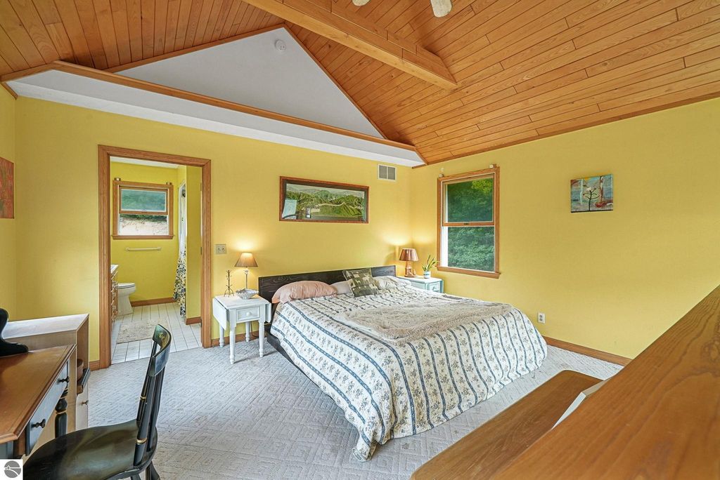 Bright yellow bedroom with vaulted wood ceiling, featuring a bed with patterned bedding, bedside tables with lamps, and a view of the surrounding woods through large windows; includes an adjoining bathroom doorway.
