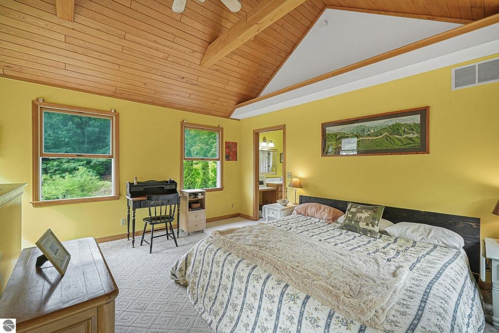Bright yellow bedroom with vaulted wood ceiling, featuring a bed with patterned bedding, two windows with views of greenery, a small desk and chair, and a nightstand with a lamp, showcasing a cozy, inviting atmosphere in a farmhouse setting near Leland, MI.