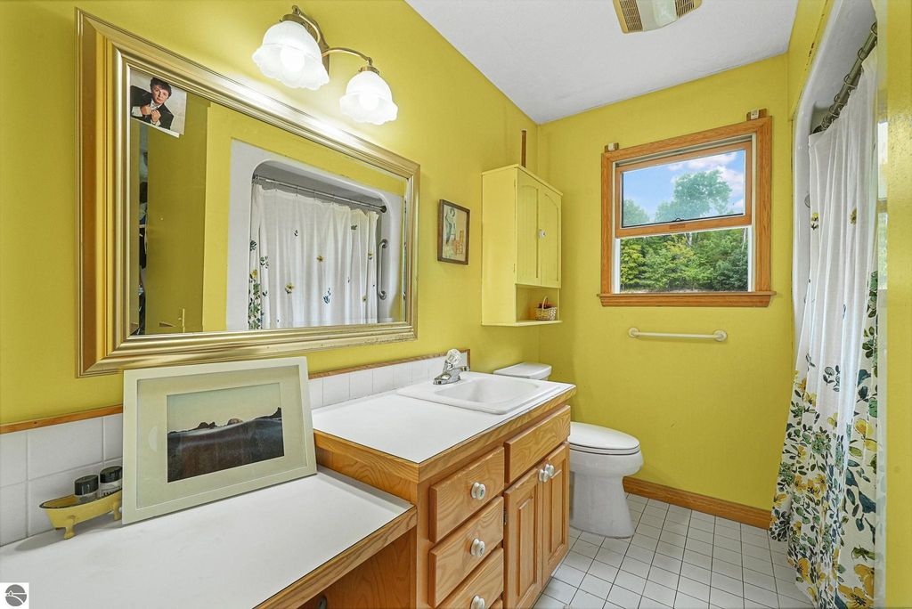 Bright yellow bathroom featuring a double sink vanity, large mirror, and a window with views of greenery, complemented by floral curtains and wooden cabinetry.