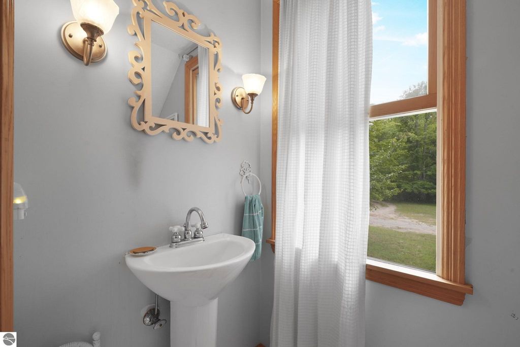 Bathroom with a white pedestal sink, decorative mirror, and natural light from a window, featuring a view of greenery outside, in a farmhouse near Leland, MI.