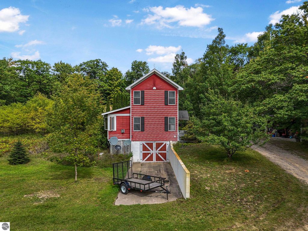 Red elevated farmhouse at 5361 N Manitou Trail, surrounded by trees and greenery, featuring a shared driveway and a trailer in front, representing a unique real estate listing in Leland, MI.