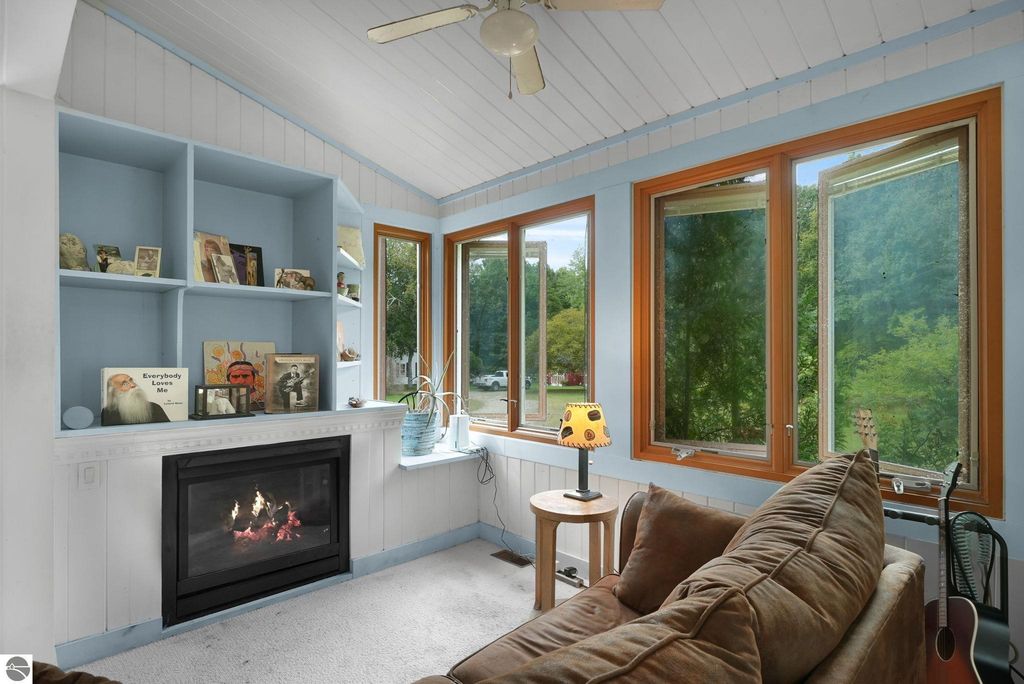 Cozy living room with a fireplace, large windows showcasing views of nature, and a comfortable sofa, featuring a light blue color scheme and decorative shelves with personal items.