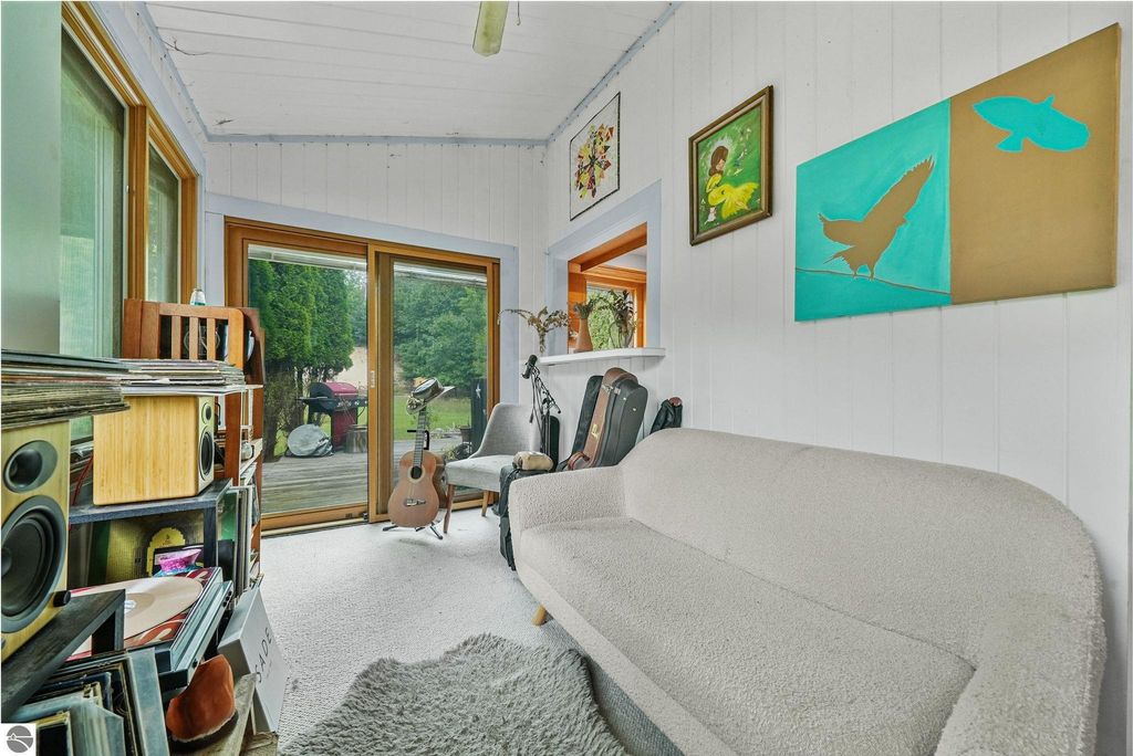 Cozy sunroom with a white sofa, wooden shelves, and a guitar, featuring large windows overlooking a deck and greenery, highlighting a relaxed atmosphere in a farmhouse near Leland, MI.