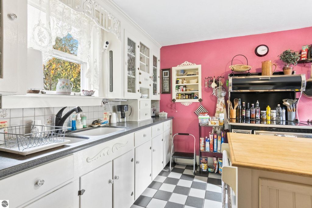 Victorian-style kitchen featuring white cabinetry, a pink accent wall, black and white checkered flooring, and modern appliances, reflecting a blend of historic charm and contemporary updates in the Onekama property listing.