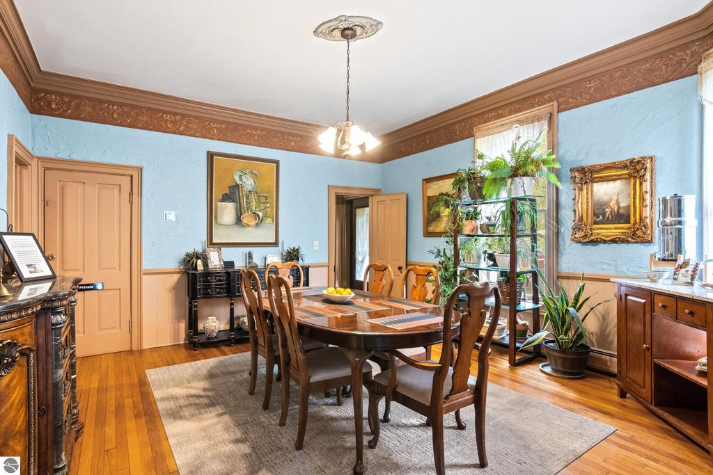 Victorian dining room featuring a large wooden table surrounded by chairs, blue walls with decorative molding, original artwork, plants, and elegant furnishings, showcasing the historic charm of the home at 8195 Fifth Street, Onekama, MI.