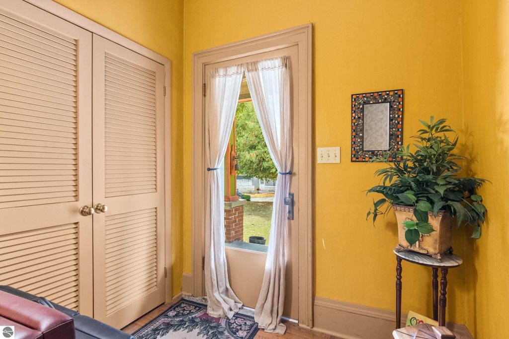 Interior view of a Victorian home entryway featuring yellow walls, closet doors, a doorway with sheer curtains leading to the outside, a decorative mirror, and a potted plant on a small table, showcasing the charm and character of the property at 8195 Fifth Street, Onekama, MI.