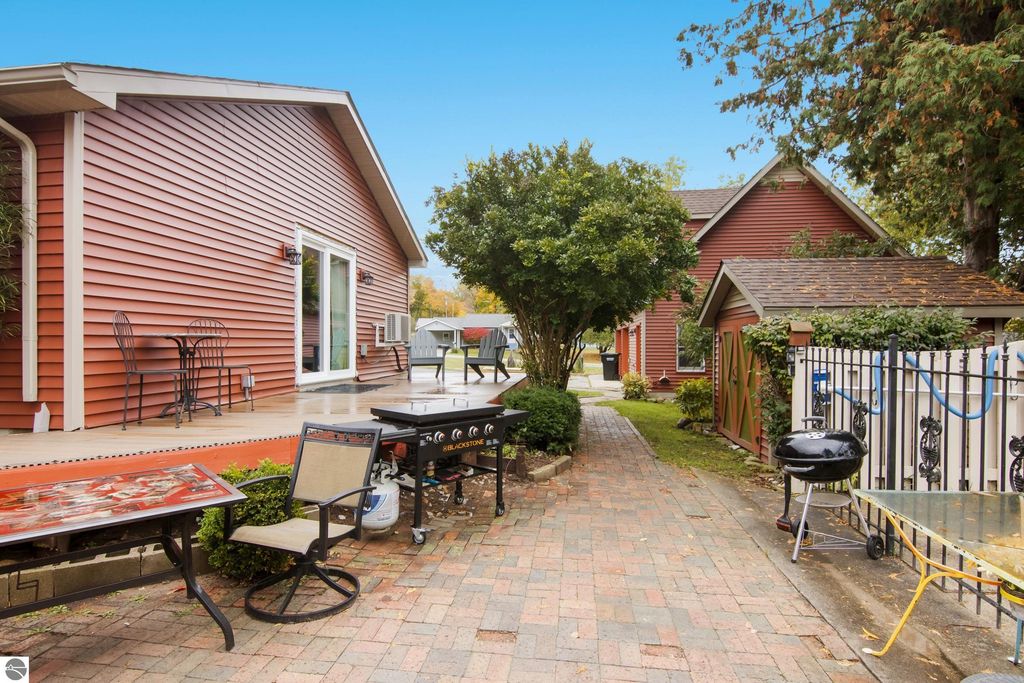 Victorian home exterior in Onekama, MI, featuring a brick pathway, outdoor seating area, and modern updates, with a glimpse of a nearby garage and lush greenery.