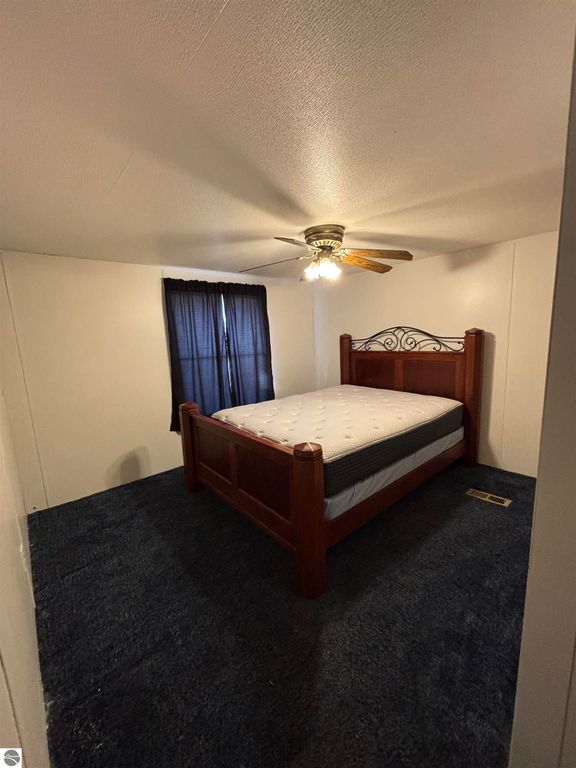 Bedroom with a wooden bed frame and mattress, ceiling fan, blue carpet, and black curtains in a manufactured home at 5013 Turner Pine Drive, Prescott, MI.