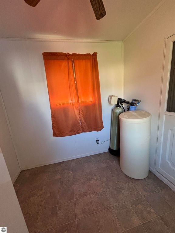 Interior view of a manufactured home featuring a window with orange curtains, a white water softener, and a gas cylinder, highlighting the home's utility space near Prescott, MI.