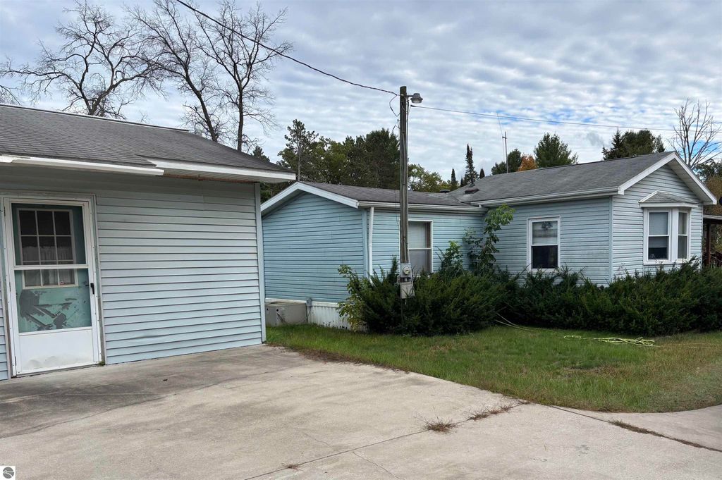Exterior view of a light blue manufactured home with an addition, featuring a concrete driveway and surrounding greenery, located at 5013 Turner Pine Drive, Prescott, MI, near lakes and amenities.