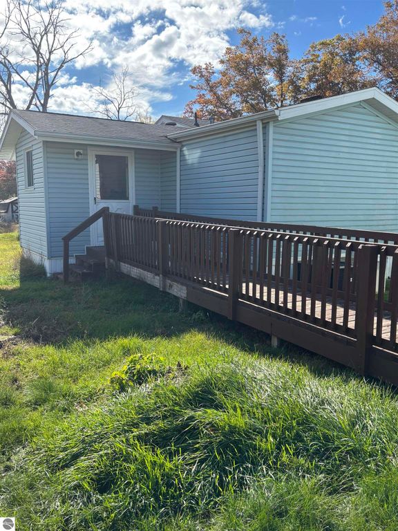 Exterior view of a 2-bedroom, 1-bath manufactured home with a wooden ramp and porch, situated on a grassy lot in Prescott, MI, highlighting its accessibility features and nearby natural surroundings.