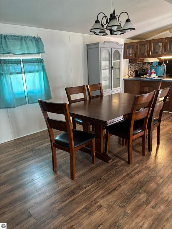 Dining area featuring a wooden table with six chairs, light blue curtains, and a glimpse of the kitchen in a manufactured home at 5013 Turner Pine Drive, Prescott, MI.