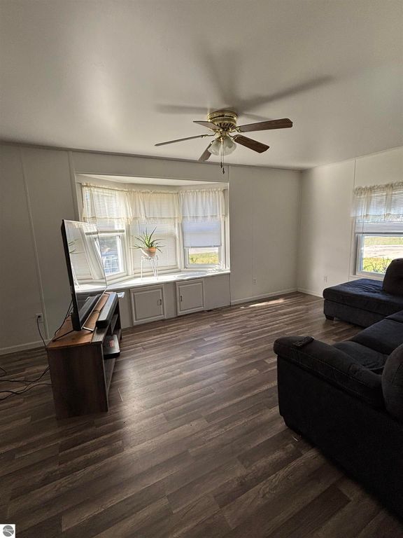 Living room of a 2-bedroom manufactured home in Prescott, MI, featuring a ceiling fan, large windows with natural light, a television on a stand, and a dark sectional sofa, showcasing the interior space and layout.