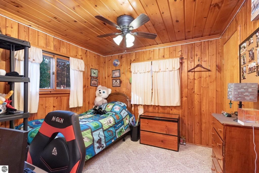 Cozy bedroom with knotty pine walls, featuring a bed with colorful bedding, a gaming chair, and a window with white curtains, highlighting a warm and inviting atmosphere.
