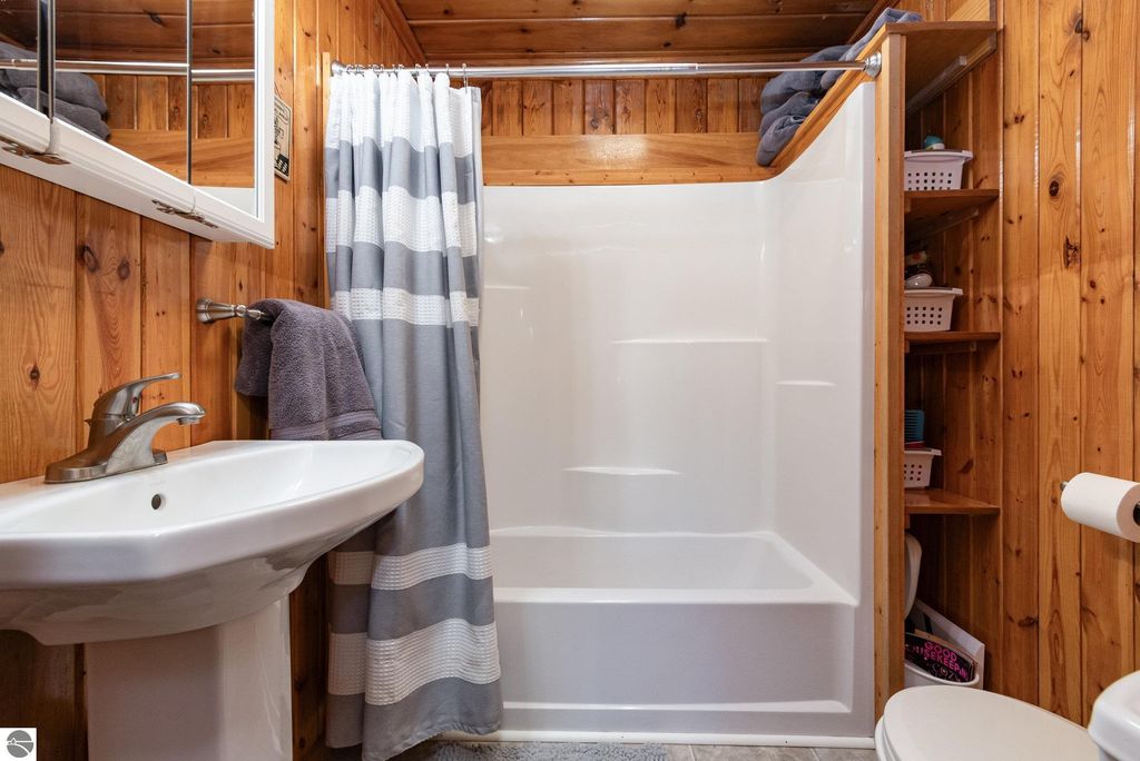 Bathroom featuring a white sink and faucet, shower with gray striped curtain, wooden paneling, and shelving with storage baskets.
