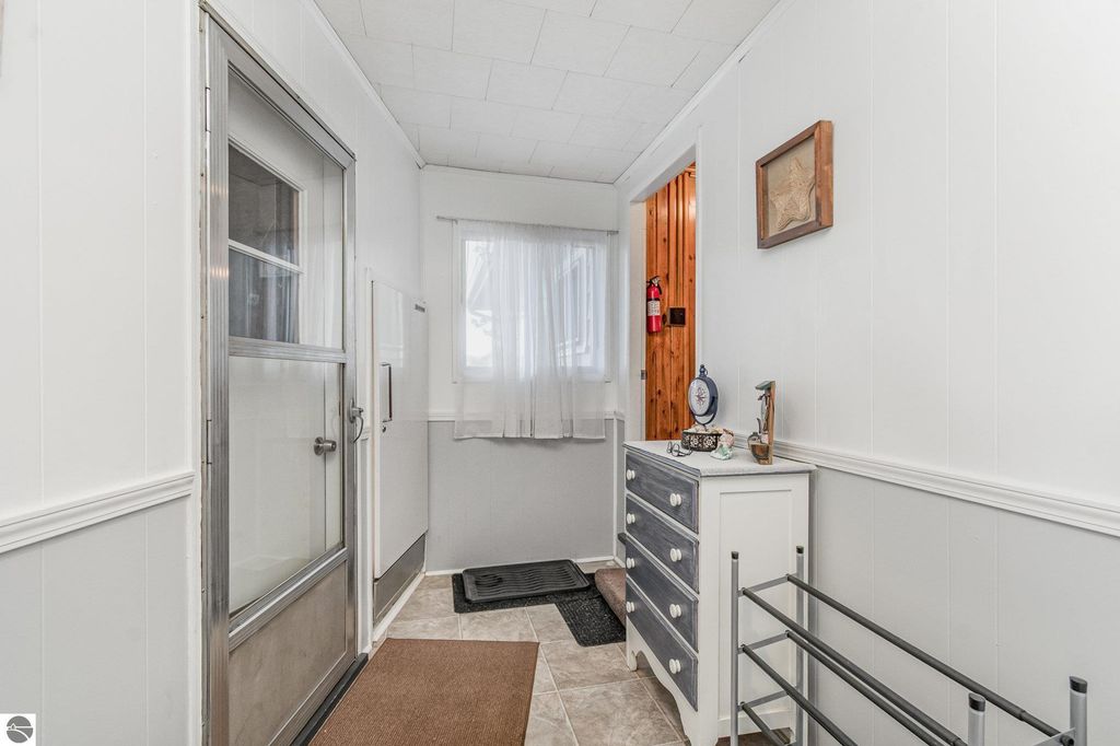 Entryway of a remodeled home featuring a glass door, white walls, a window with sheer curtains, a decorative wall frame, a dresser, and a shoe rack, highlighting the welcoming atmosphere of the property at 2096 Timber Trail, National City, MI.