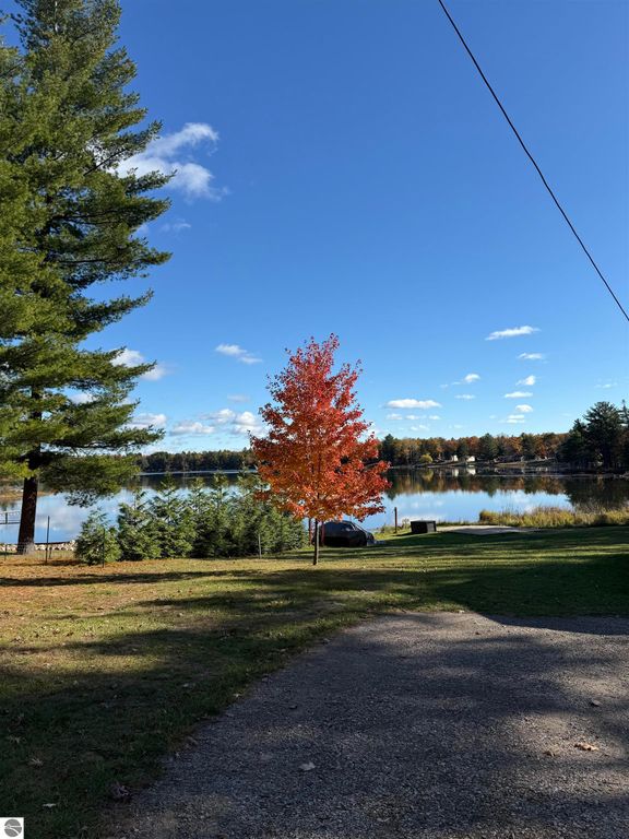 Scenic view of Big Island Lake with vibrant autumn foliage, featuring a red maple tree, green pine trees, and a clear blue sky, highlighting the tranquil waterfront setting near 2096 Timber Trail, National City, MI.
