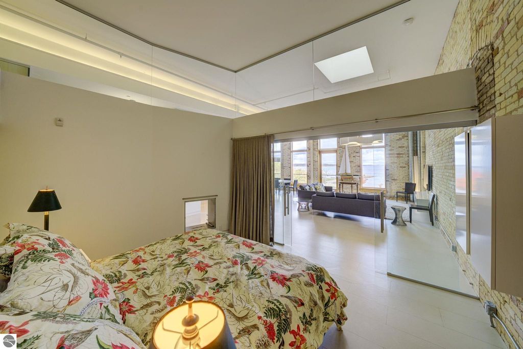 Modern bedroom with floral bedding, glass walls, and views of a spacious living area featuring large windows, showcasing the contemporary design of a downtown Traverse City condo.