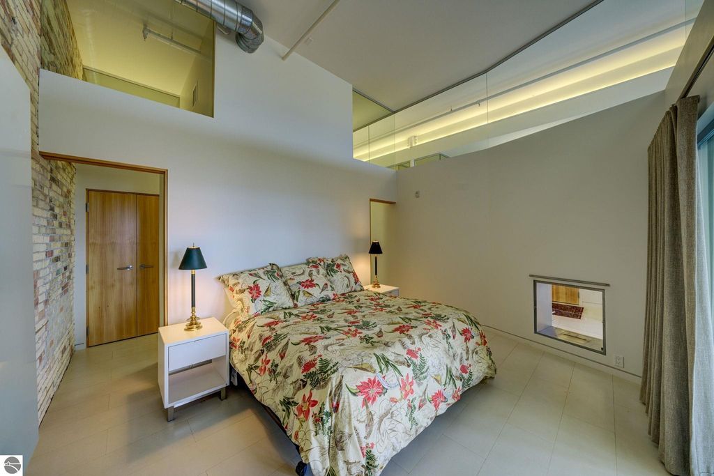 Modern bedroom in downtown Traverse City condo, featuring floral bedding, bedside lamp, and large windows, highlighting contemporary design and spacious layout.