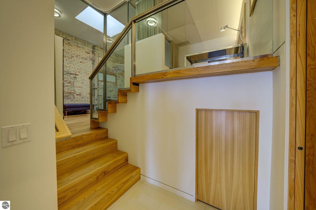 Modern interior staircase with wooden steps and glass railing, leading to a bright space with exposed brick walls, reflecting contemporary design elements of the downtown condo at 237 E Front Street, Traverse City.