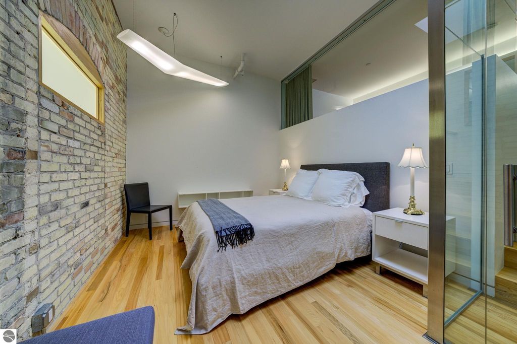 Modern bedroom featuring a queen-sized bed, minimalist decor, exposed brick wall, wooden flooring, and stylish lighting, located in a downtown Traverse City condo.