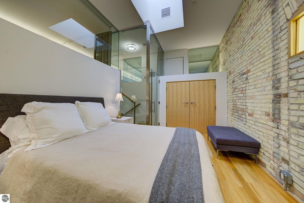 Modern bedroom in downtown Traverse City condo, featuring exposed brick walls, large windows, and stylish furnishings, emphasizing contemporary design and spaciousness.
