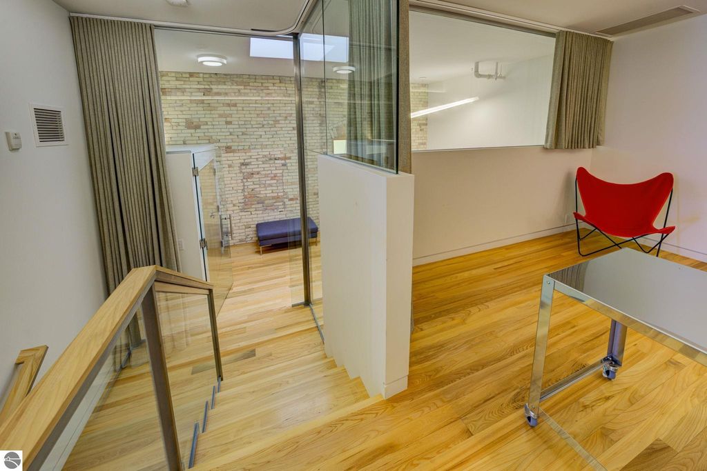Modern condo interior featuring wooden floors, glass walls, and a red accent chair, showcasing a spacious layout and natural light, ideal for downtown living in Traverse City.