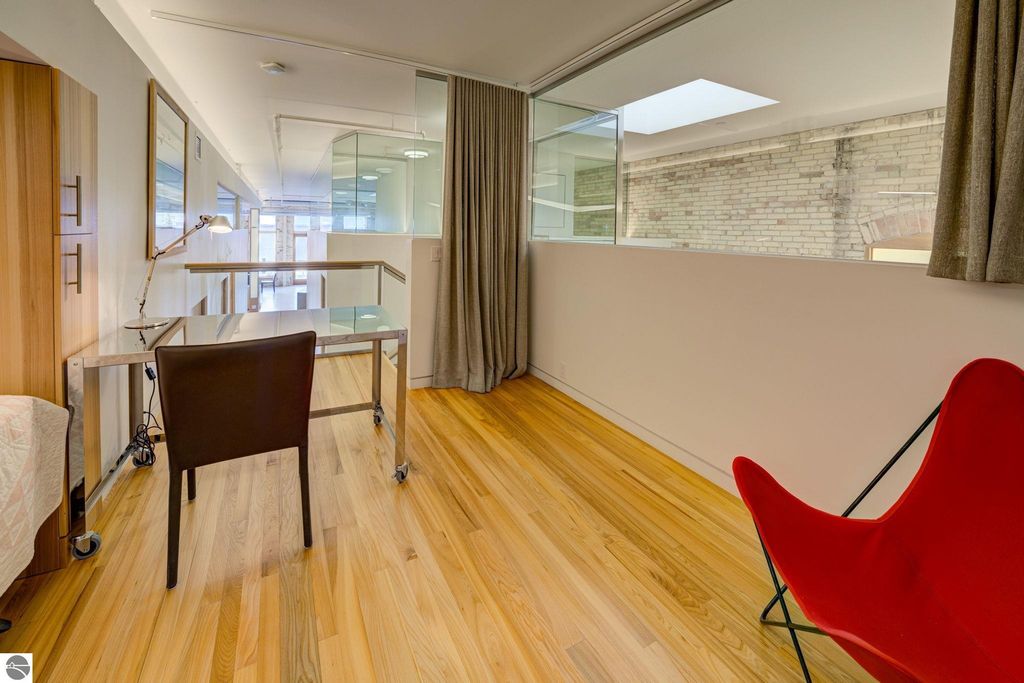 Modern downtown condo interior featuring a sleek glass desk, brown leather chair, and vibrant red accent chair, complemented by wooden flooring and natural light from skylights, showcasing contemporary design elements.