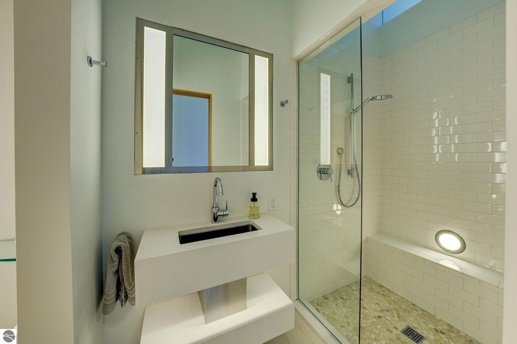 Modern bathroom featuring a sleek vanity with a minimalist sink, large mirror, glass shower enclosure with rainfall showerhead, and pebble stone flooring, reflecting the contemporary design of the downtown condo at 237 E Front Street, Traverse City, MI.
