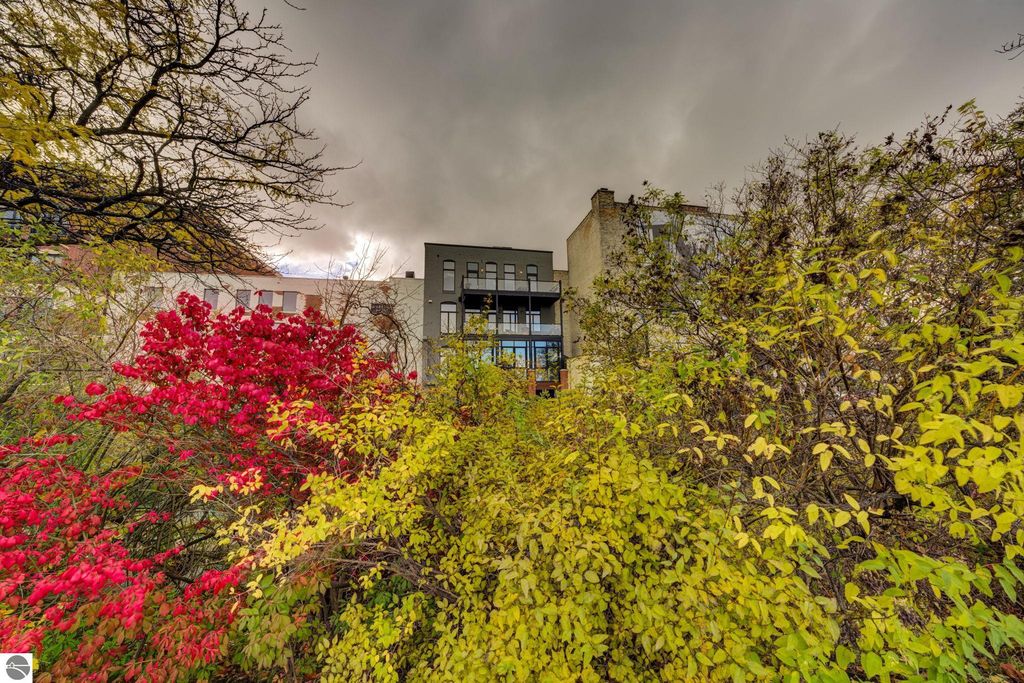 Modern downtown condo at 237 E Front Street, Traverse City, framed by vibrant autumn foliage, showcasing large windows and balcony, with a backdrop of a dramatic sky.