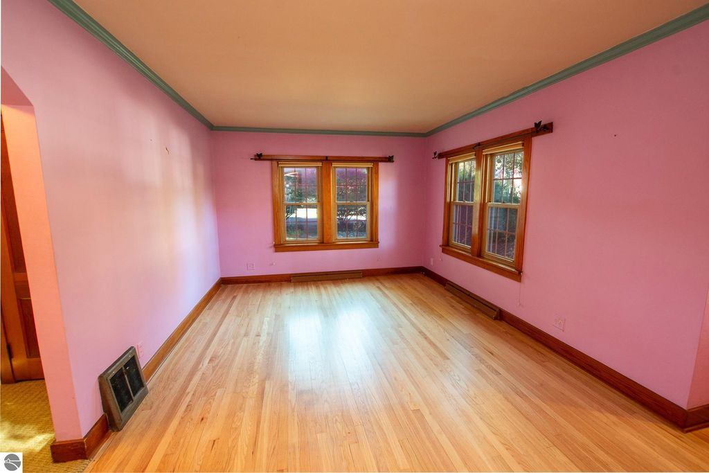 Interior view of a vacant room with pink walls and hardwood flooring, featuring large windows with wooden trim, showcasing natural light and potential for renovation.