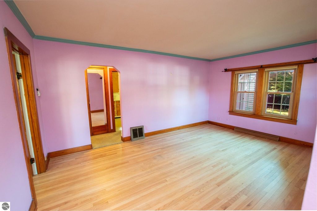 Interior view of a vacant room featuring light purple walls, hardwood flooring, and a large window, showcasing potential for renovation in the 2-bedroom ranch home at 1101 Jefferson Avenue, Traverse City, MI.
