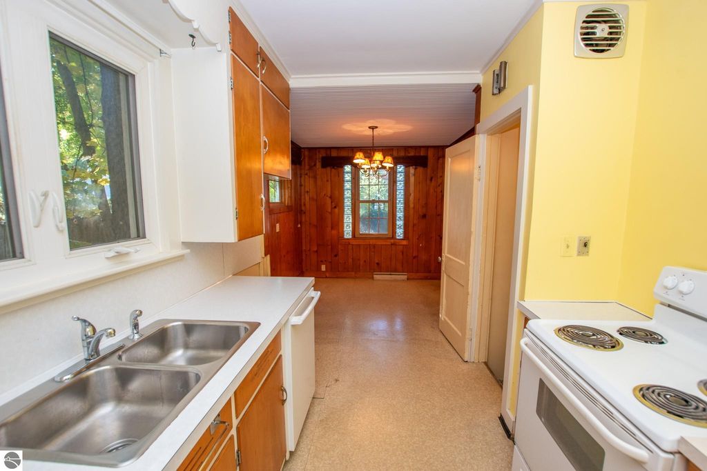 Kitchen interior featuring stainless steel double sink, wooden cabinetry, and a view into a dining area with wood paneling and chandelier, showcasing the potential of the 2-bedroom ranch home at 1101 Jefferson Avenue, Traverse City, MI.