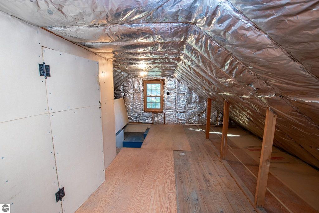 Attic space featuring exposed wooden flooring, silver insulation on the sloped ceiling, and a window providing natural light, ideal for renovation or conversion in Traverse City home listing.
