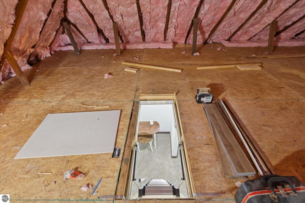 Attic space with pink insulation, wooden beams, and a hatch leading to a room with a chair, showcasing the updated indoor storage area of the renovated 3-bedroom home at 117 E Wright Avenue, Shepherd, MI.