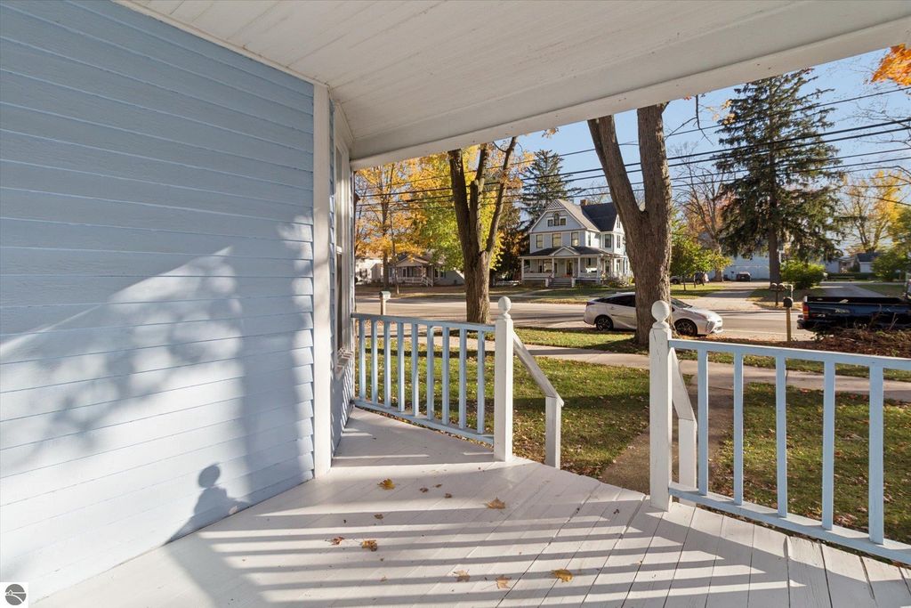 Porch view of a renovated home at 117 E Wright Avenue in Shepherd, MI, featuring a light blue exterior and white railing, surrounded by trees and residential street.